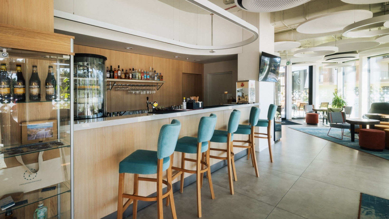 Nowoczesny bar hotelowy z jasnym drewnem, turkusowymi stołkami barowymi i jasnym, przestronnym wnętrzem z dużymi oknami.