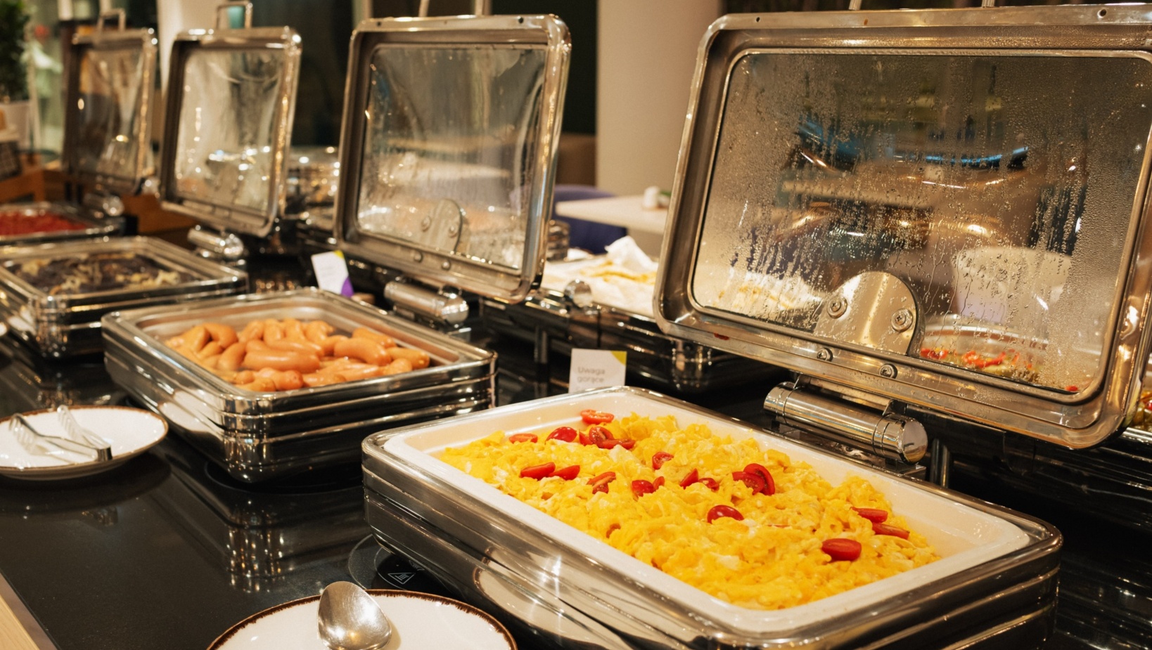 Bufet śniadaniowy w hotelu z podgrzewanymi potrawami: jajecznica z pomidorami i parówki w stalowych pojemnikach.