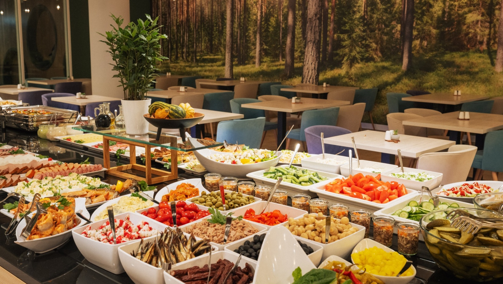Bufet restauracyjny z różnorodnymi sałatkami, świeżymi warzywami i przystawkami w jadalni z motywem leśnym na ścianie.