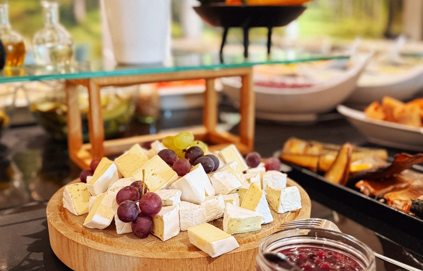 Deska serów z różnorodnym serem i winogronami, obok słoik owocowego dżemu na bufecie śniadaniowym w hotelu.