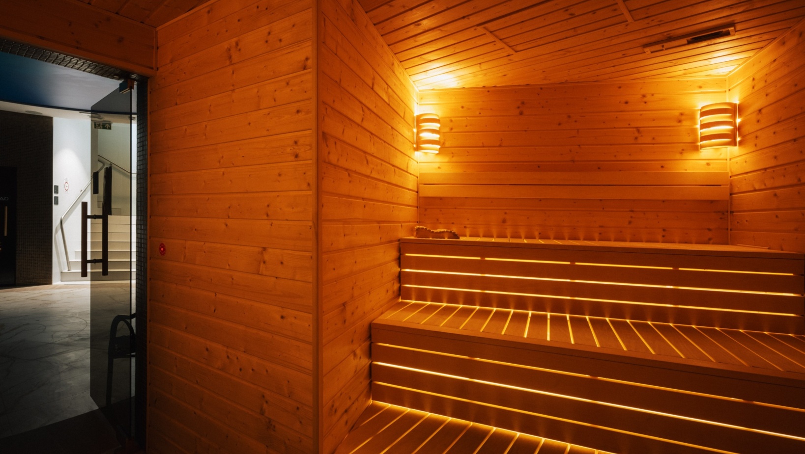 Przytulna sauna hotelowa z drewnianymi ławami podświetlanymi ciepłym światłem i nowoczesnym oświetleniem ściennym.