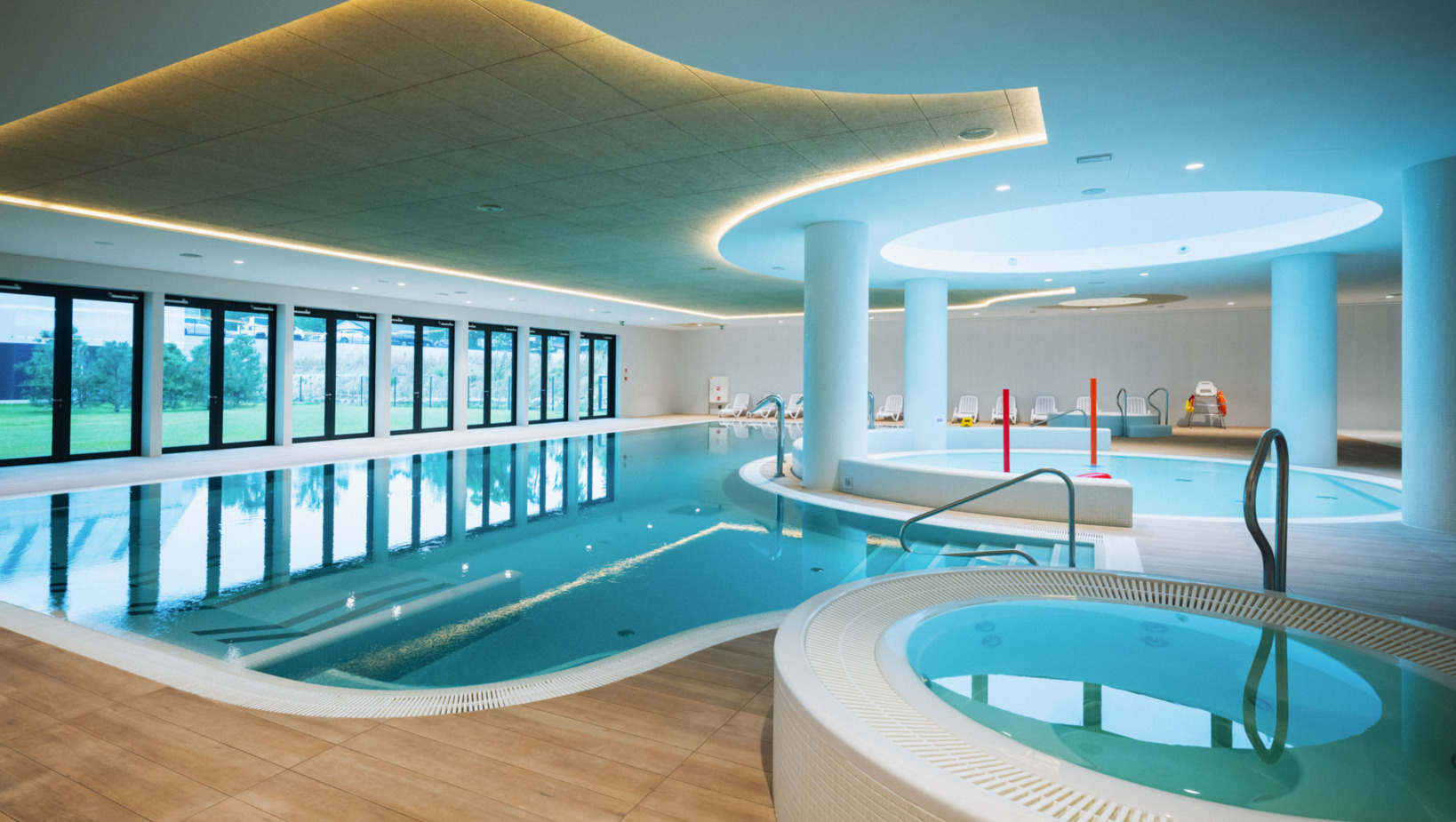 Przestronny, nowoczesny basen kryty z jacuzzi, leżakami i dużymi oknami z widokiem na zielony ogród.