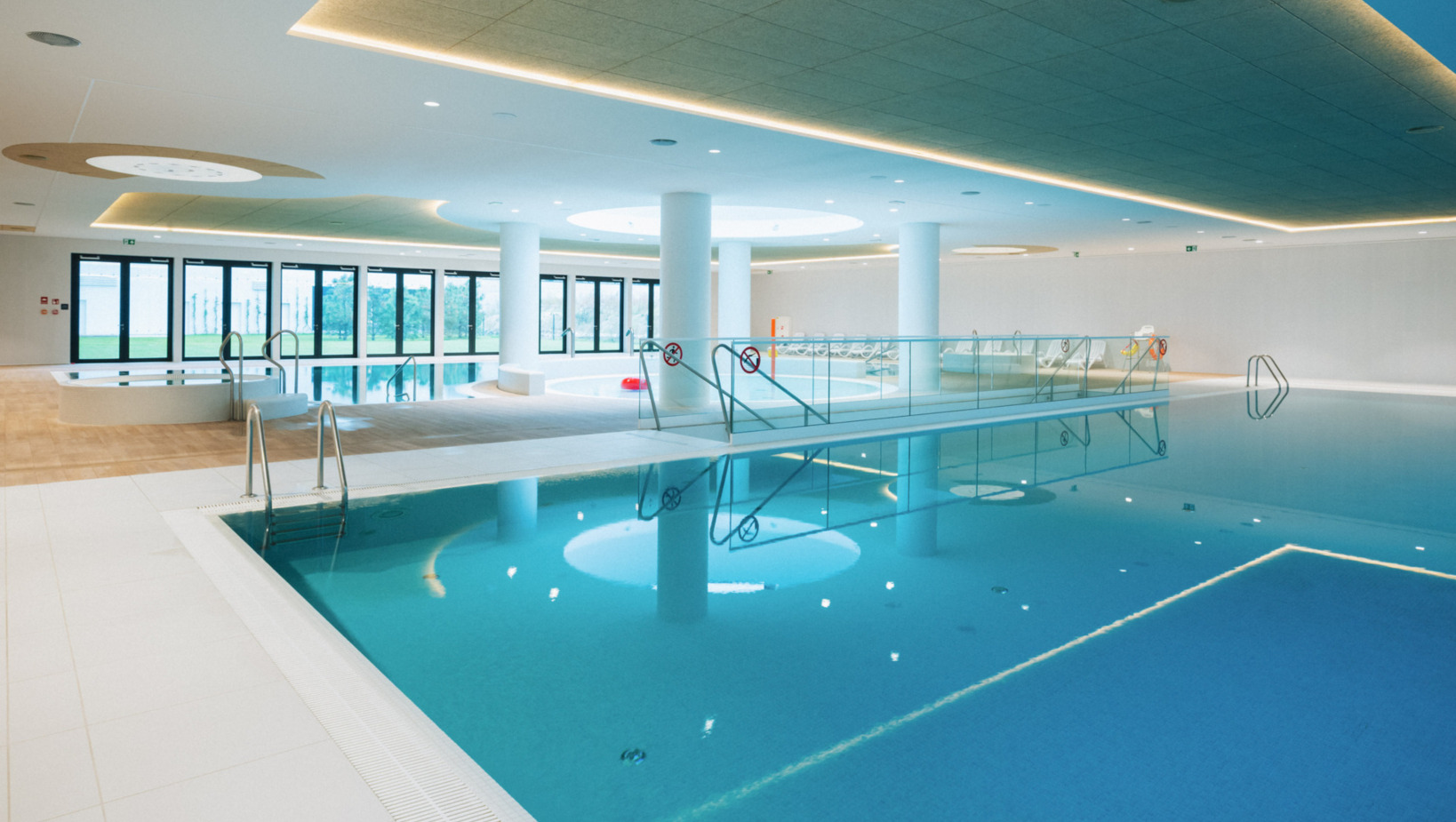 Przestronny, nowoczesny basen kryty z oświetleniem LED i panoramicznymi oknami z widokiem na zieleń na zewnątrz.