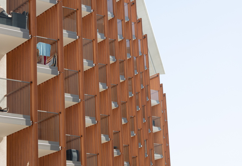 Nowoczesny hotel z balkonami wyposażonymi w przesłony z drewna i przeszklone balustrady, otoczony zielenią.