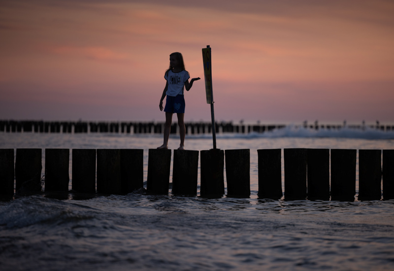 Dziecko balansujące na drewnianych palach przy brzegu morza o zmierzchu z różowym niebem w tle