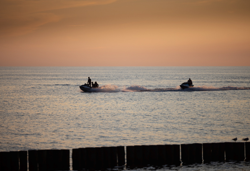 Goście na skutrach wodnych pływają po spokojnym morzu o zachodzie słońca, widok z plaży z drewnianymi palami.