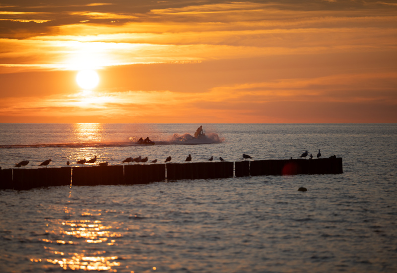 Zachód słońca nad morzem z sylwetkami osób na skuterze wodnym i ptakami na drewnianych falochronach.