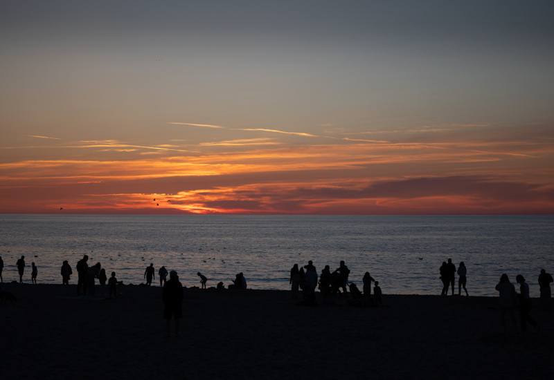 Tłum ludzi na plaży o zmierzchu, podziwiających kolorowy zachód słońca nad spokojnym morzem