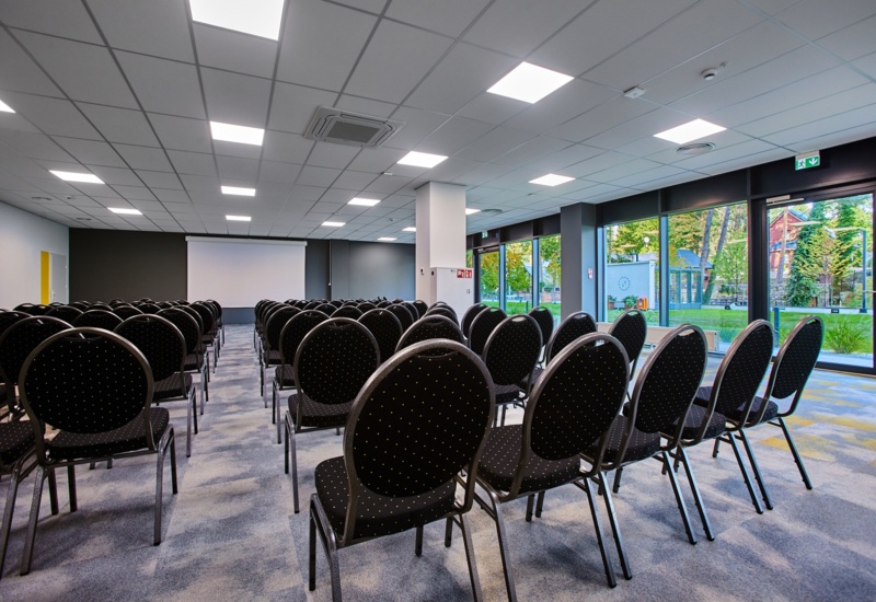 Nowoczesna sala konferencyjna z rzędem czarnych krzeseł, projektorem i dużymi oknami z widokiem na ogród.