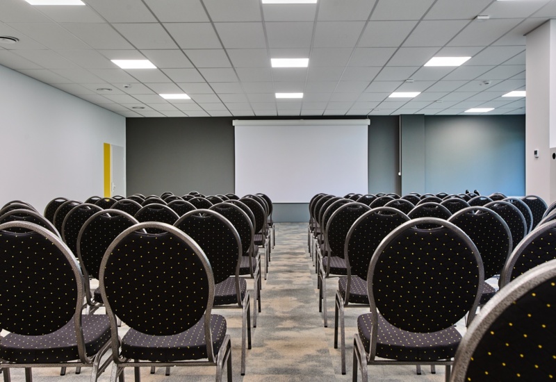 Nowoczesna sala konferencyjna z rzędami czarnych krzeseł i dużym ekranem projekcyjnym na szarej ścianie