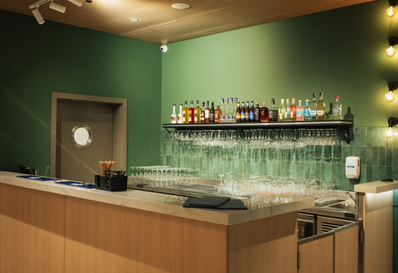 Nowoczesny hotelowy bar z zielonymi ścianami, drewnianym blatem i półką z alkoholem oraz kieliszkami.