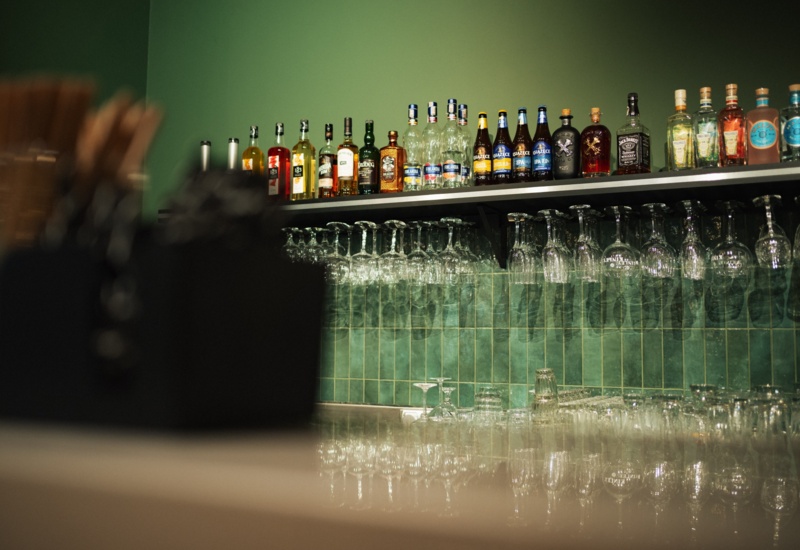 Półka hotelowego baru z różnorodnymi butelkami alkoholu i wiszącymi kieliszkami na tle zielonych płytek.