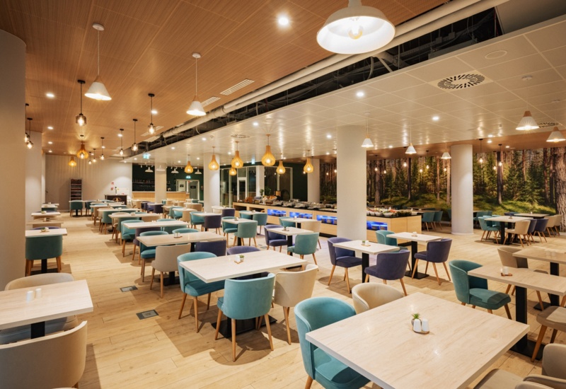 Elegancka restauracja hotelowa z jasnym drewnianym wnętrzem, miękkimi fotelami i bufetem z dekoracjami roślinnymi.