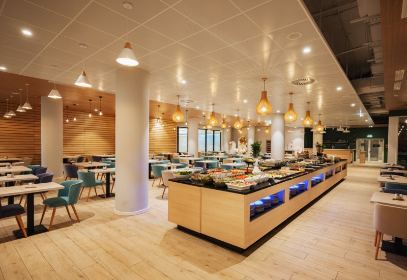 Nowoczesna hotelowa jadalnia z bufetem pełnym świeżych potraw i eleganckimi, kolorowymi krzesłami przy drewnianych stołach.