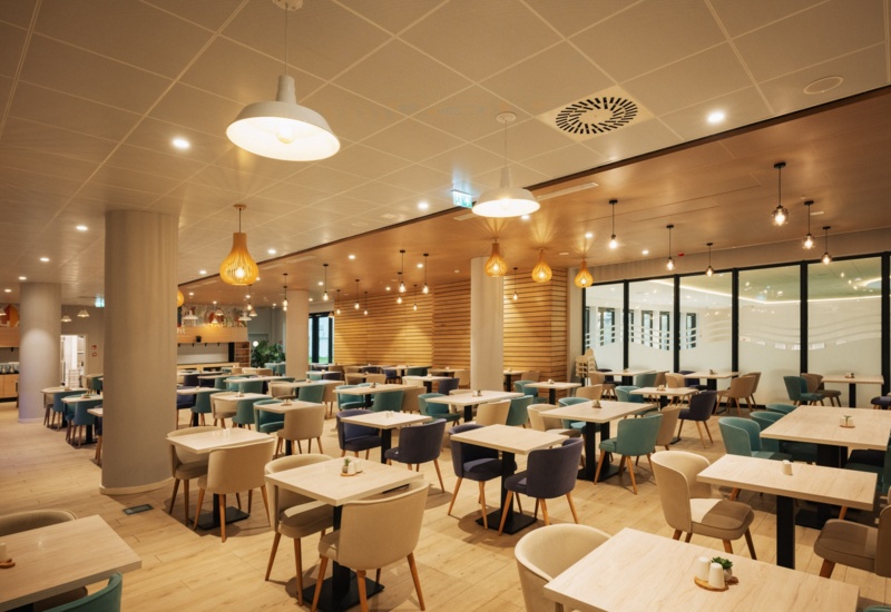 Przestronna nowoczesna restauracja hotelowa z drewnianymi stołami, kolorowymi wygodnymi fotelami i ciepłym oświetleniem.