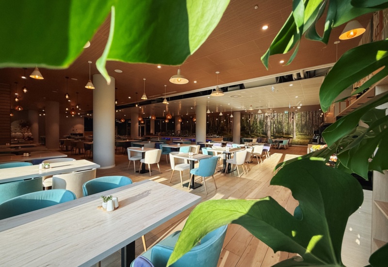 Jasna, przestronna restauracja hotelowa z drewnianymi stołami, miękkimi turkusowymi i kremowymi krzesłami, dekoracją ścienną z lasem.