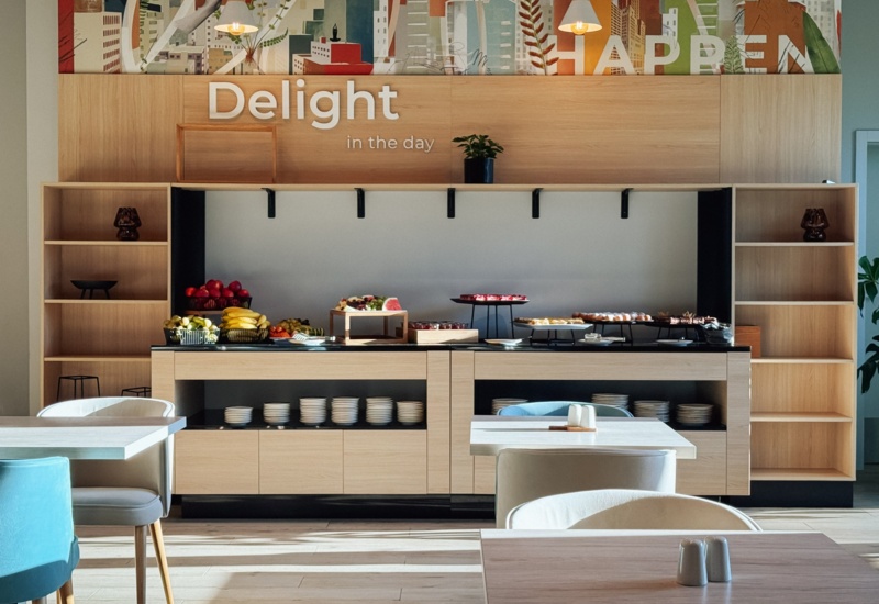 Jasna jadalnia z bufetem oferującym świeże owoce i słodkości, nowoczesne meble i dekoracje ścienne z napisem Delight