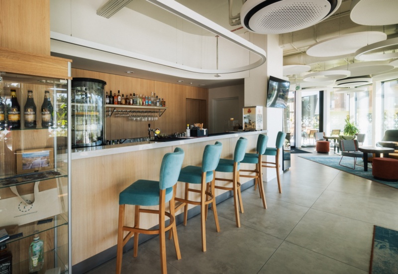 Nowoczesny bar hotelowy z jasnym drewnem, turkusowymi stołkami barowymi i jasnym, przestronnym wnętrzem z dużymi oknami.
