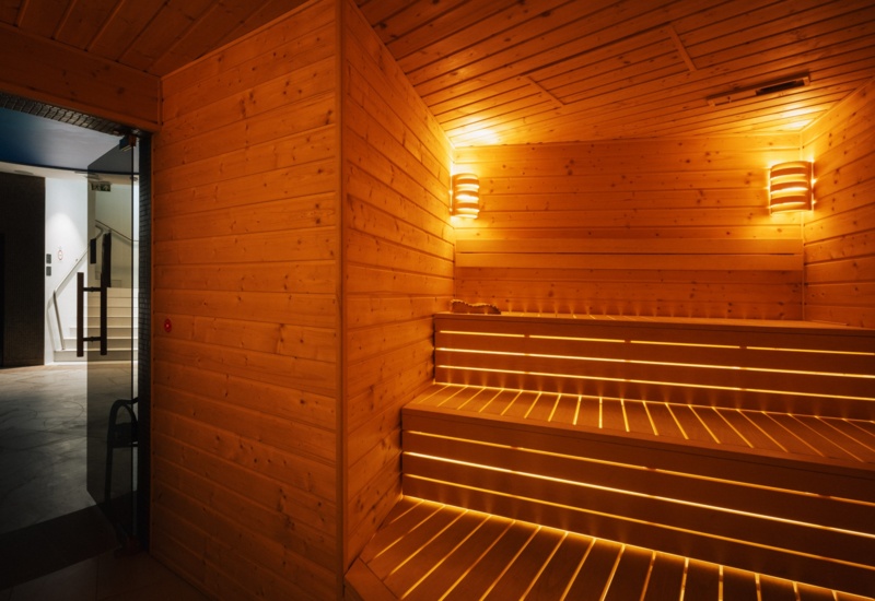 Przytulna sauna hotelowa z drewnianymi ławami podświetlanymi ciepłym światłem i nowoczesnym oświetleniem ściennym.