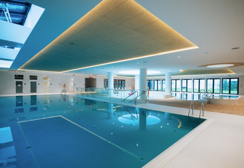 Nowoczesny kryty basen hotelowy z oświetleniem sufitowym, leżakami i dużymi oknami z widokiem na zielony ogród.