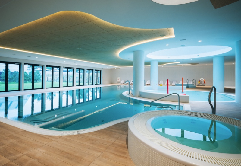 Nowoczesny kryty basen hotelowy z jacuzzi, dużymi oknami i relaksującymi leżakami przy basenie.