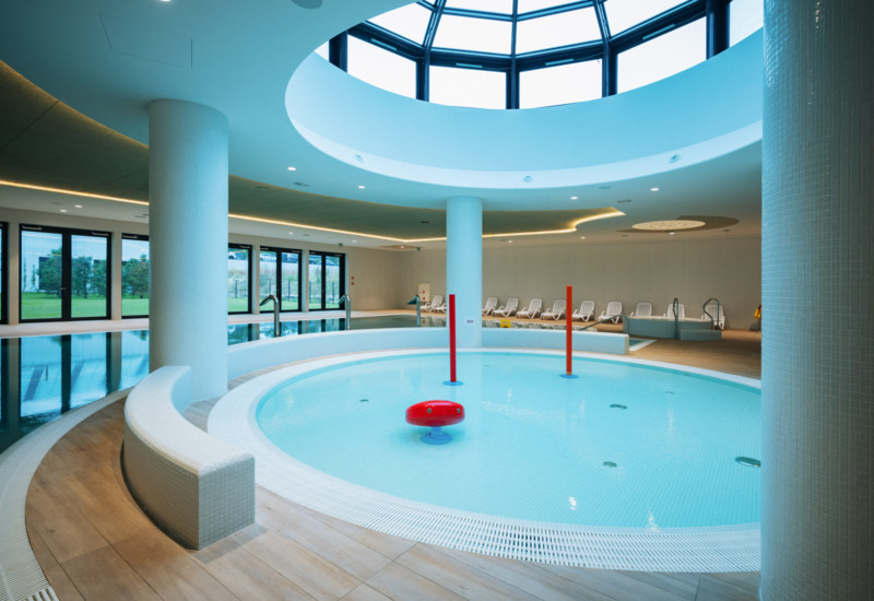 Nowoczesny hotelowy basen kryty z okrągłą niecką dla dzieci i panoramicznym oknem w suficie, leżaki w tle