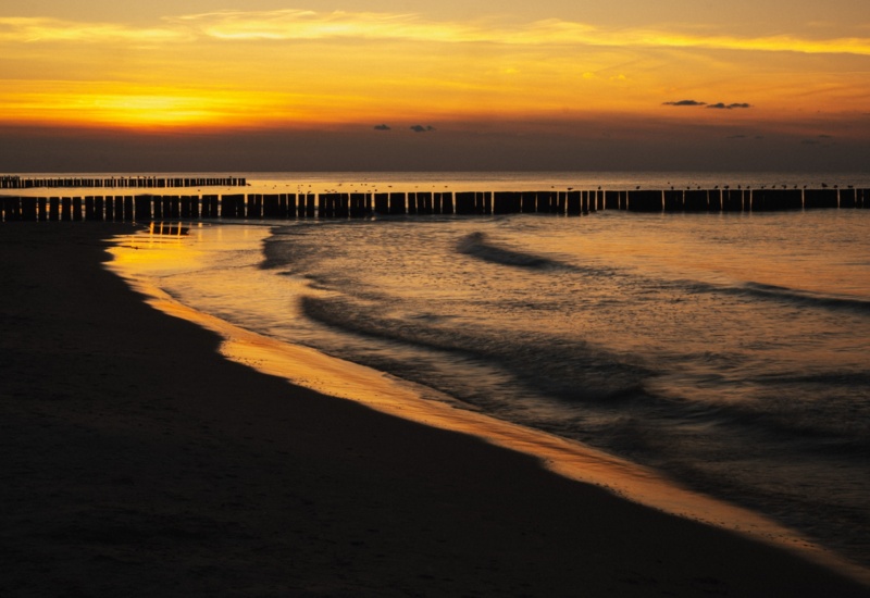 Spokojna plaża o zmierzchu z drewnianymi falochronami i delikatnie falującą wodą pod złotym niebem