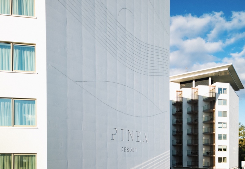 Nowoczesny budynek resortu Pinea z balkonami i dużymi oknami, na tle niebieskiego nieba z chmurami.
