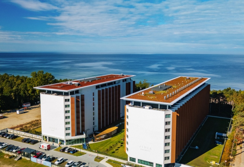 Nowoczesny kompleks apartamentów przy plaży z widokiem na morze, dużą zielenią i parkingiem dla gości.