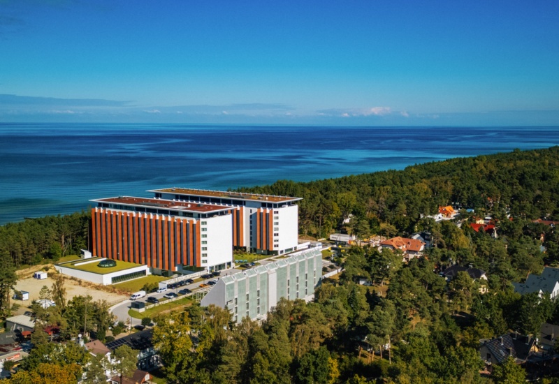 Nowoczesny hotel z czerwono-białą elewacją przy lesie, z widokiem na spokojne wybrzeże i morze.