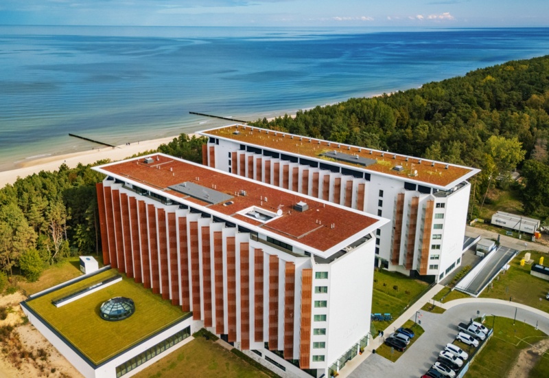 Nowoczesny hotel nadmorski z białymi budynkami, tarasami i panoramicznym widokiem na plażę i las sosnowy.