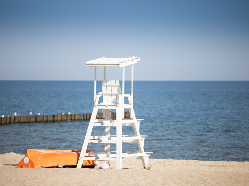Biała drewniana wieża ratownika na piaszczystej plaży z widokiem na spokojne morze i drewniany falochron.