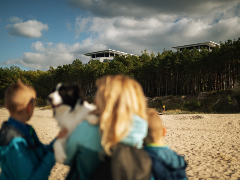 Rodzina z psem na piaszczystej plaży z widokiem na nowoczesne budynki nad lasem sosnowym pod pochmurnym niebem