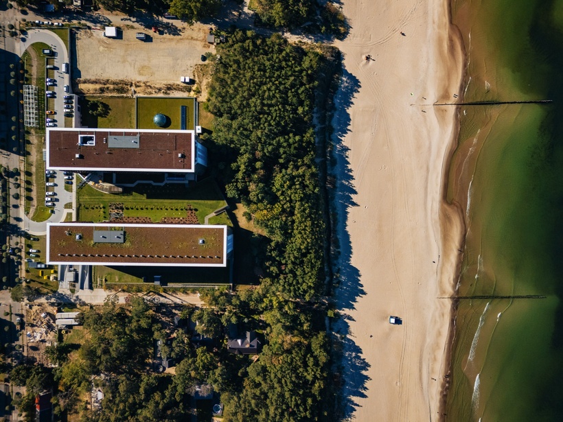 Luftaufnahme eines modernen Hotels am Strand, umgeben von Bäumen und Parkplätzen, angrenzend an einen breiten Sandstrand