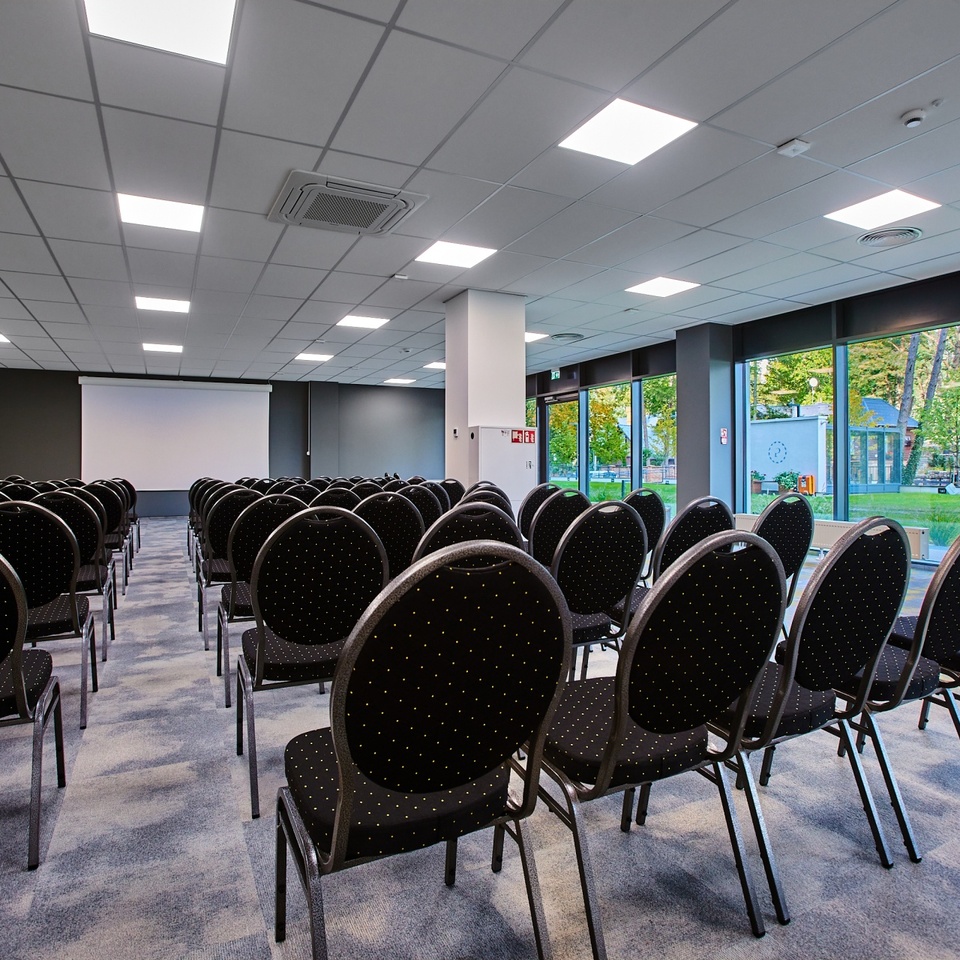 Moderner Konferenzraum mit Reihen schwarzer Stühle, Projektionsleinwand und großen Fenstern mit Gartenblick.