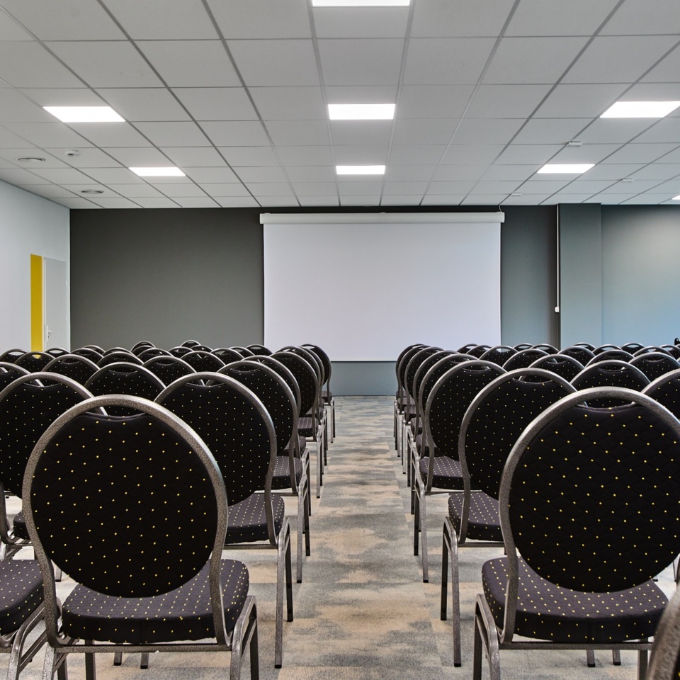 Nowoczesna sala konferencyjna z rzędami czarnych krzeseł i dużym ekranem projekcyjnym na szarej ścianie
