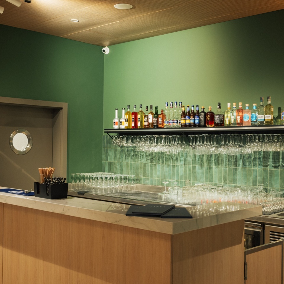 Nowoczesny hotelowy bar z zielonymi ścianami, drewnianym blatem i półką z alkoholem oraz kieliszkami.