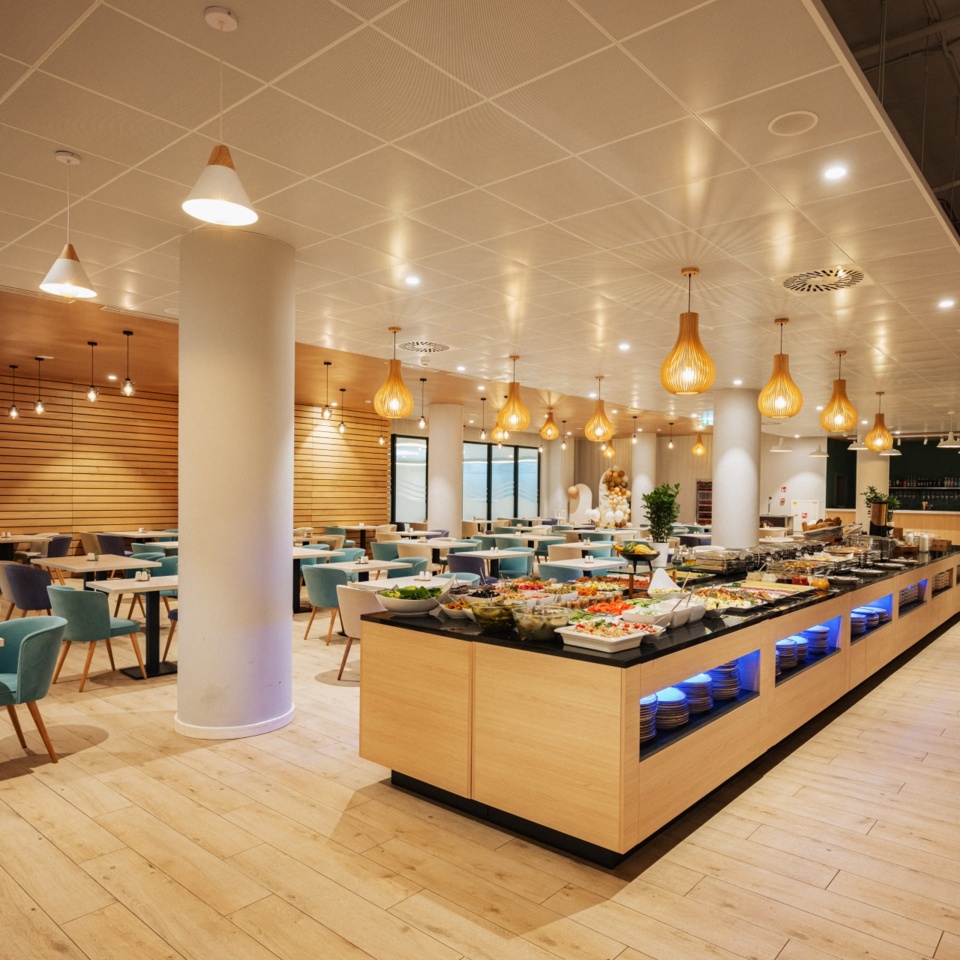 Nowoczesna hotelowa jadalnia z bufetem pełnym świeżych potraw i eleganckimi, kolorowymi krzesłami przy drewnianych stołach.