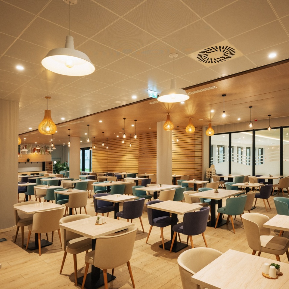 Przestronna nowoczesna restauracja hotelowa z drewnianymi stołami, kolorowymi wygodnymi fotelami i ciepłym oświetleniem.