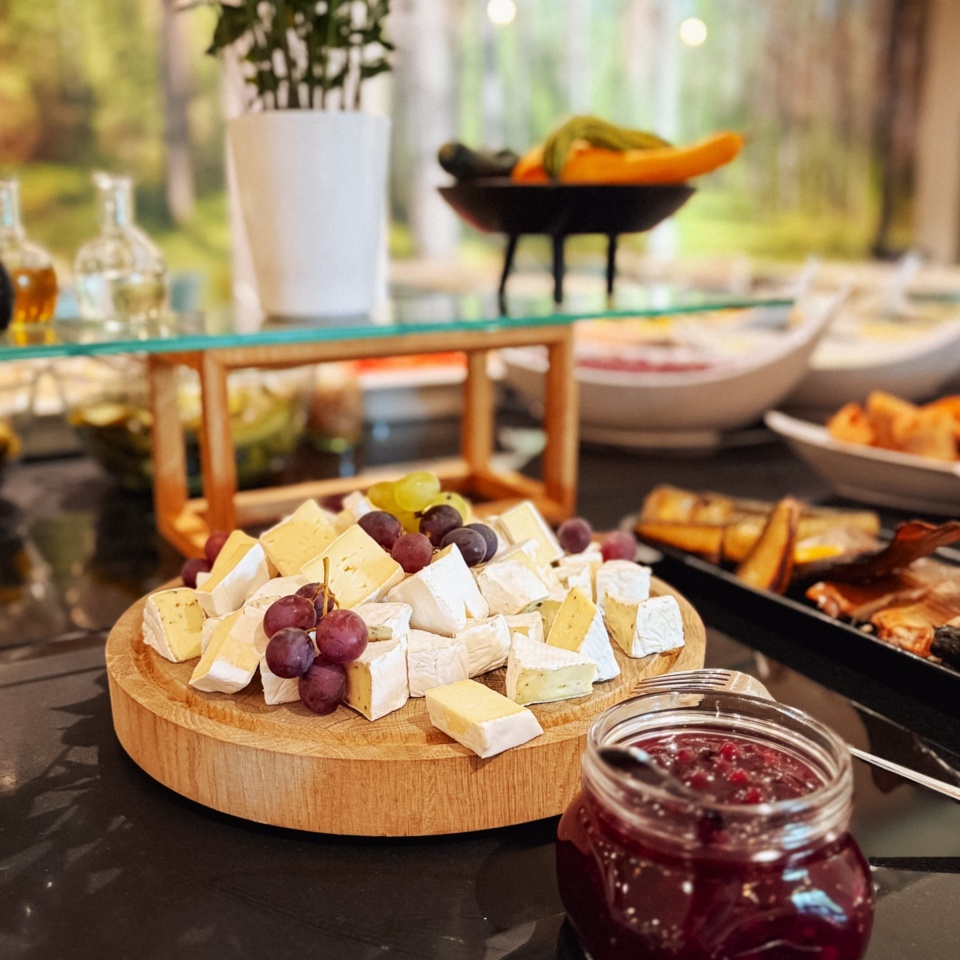 Deska serów z różnorodnym serem i winogronami, obok słoik owocowego dżemu na bufecie śniadaniowym w hotelu.