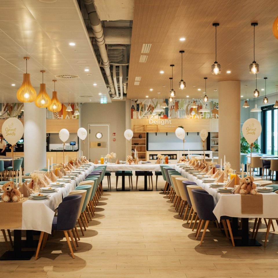 Nowoczesna sala bankietowa z długimi stołami ustawionymi na uroczystość, jasne wnętrze z drewnianymi akcentami i dekoracjami.