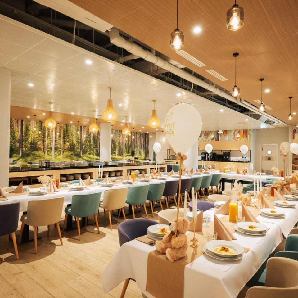 Przestronna sala bankietowa z długimi stołami, pastelowymi krzesłami, balonami i przytulną dekoracją z pluszakami.