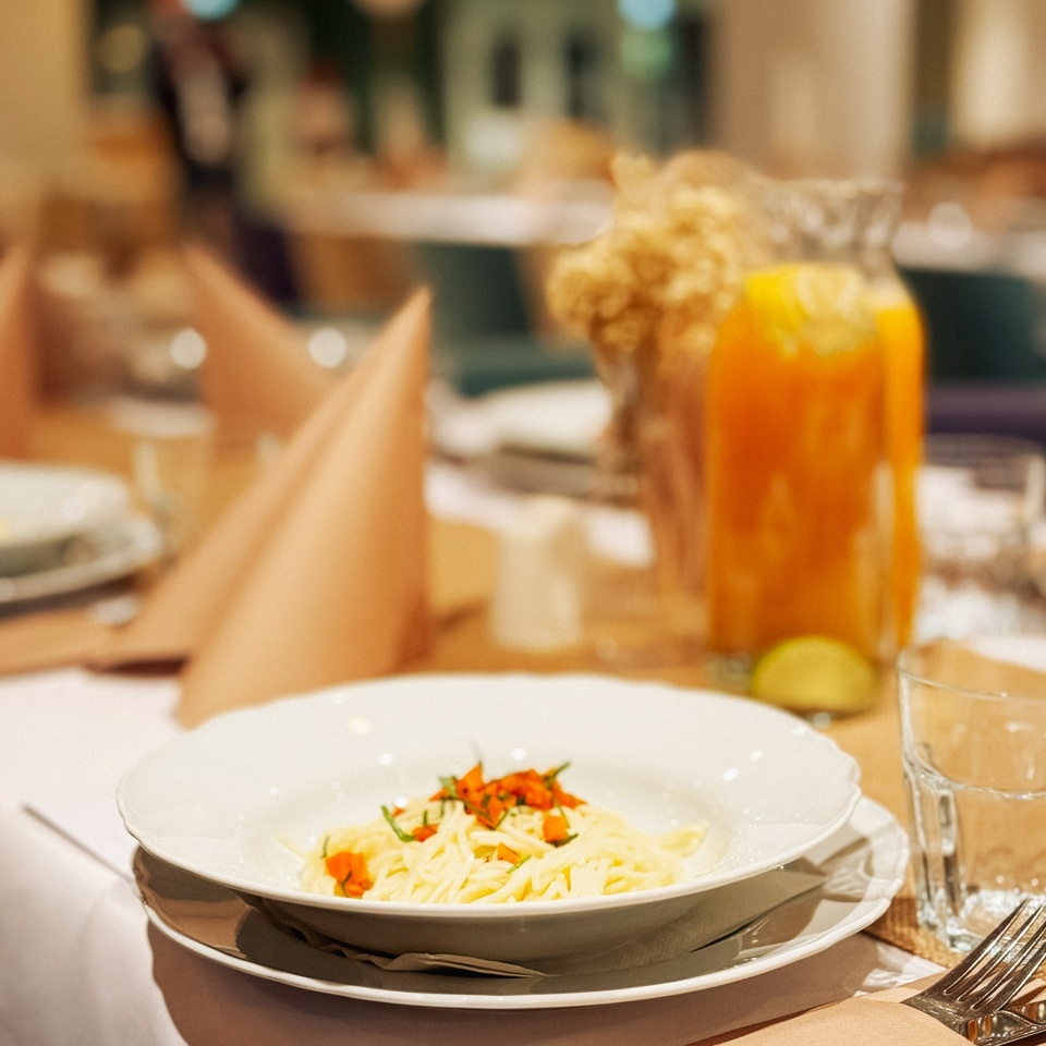 Spaghetti with herb garnish in a white bowl at an elegantly set hotel restaurant table, with a carafe of juice nearby