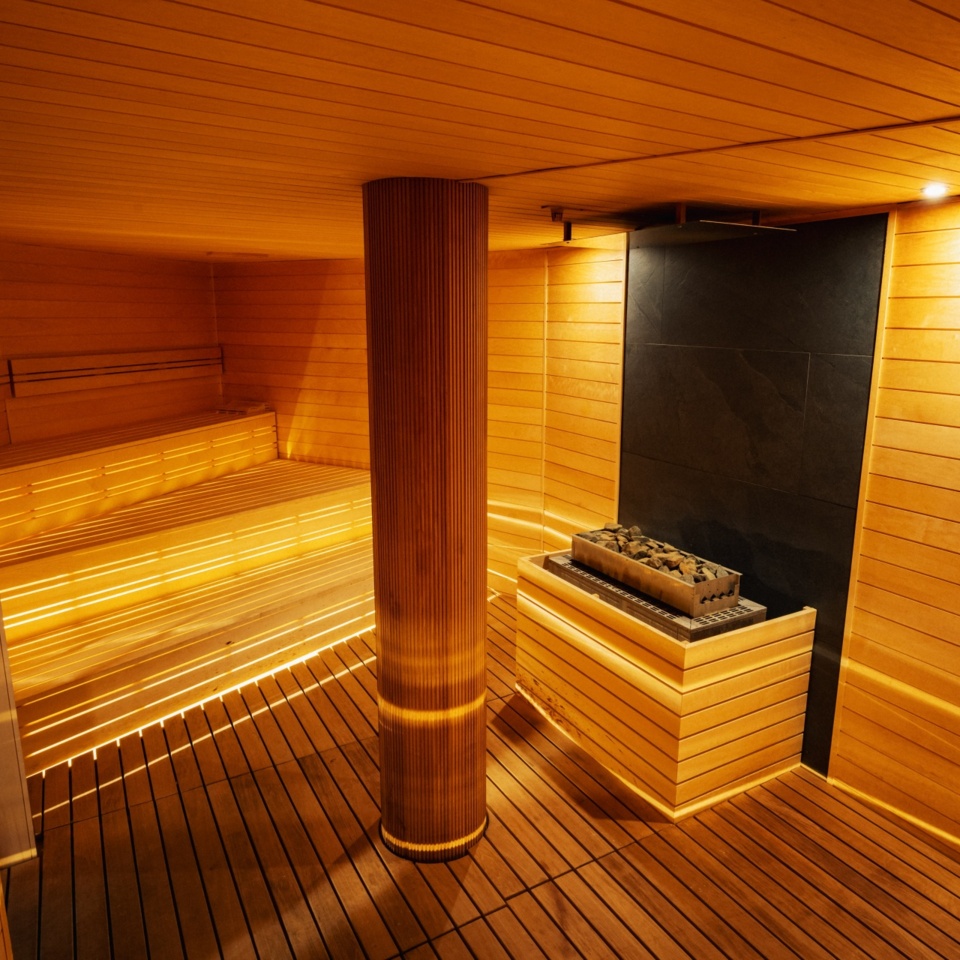 Przytulna sauna hotelowa z drewnianymi ławkami, centralną kolumną i podświetlanymi panelami ściennymi i podłogowymi.