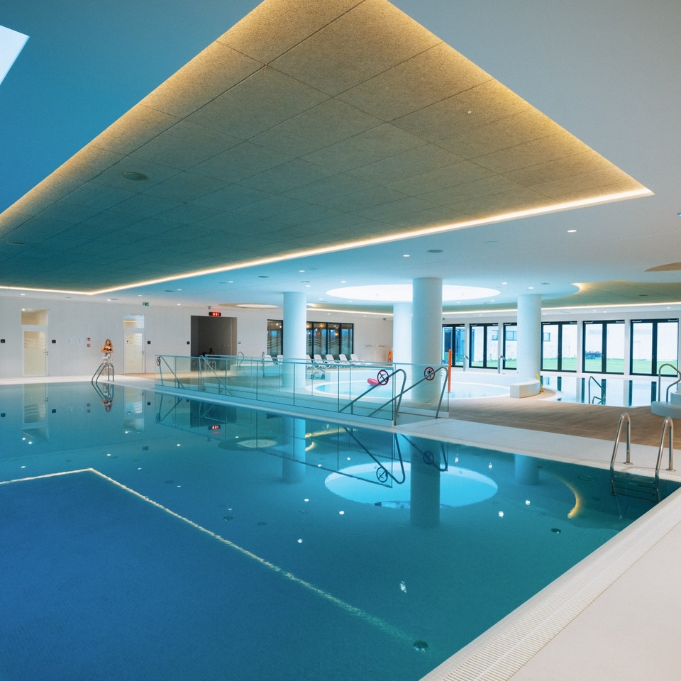 Nowoczesny kryty basen hotelowy z oświetleniem sufitowym, leżakami i dużymi oknami z widokiem na zielony ogród.