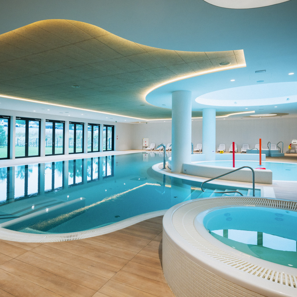 Przestronny, nowoczesny basen kryty z jacuzzi, leżakami i dużymi oknami z widokiem na zielony ogród.