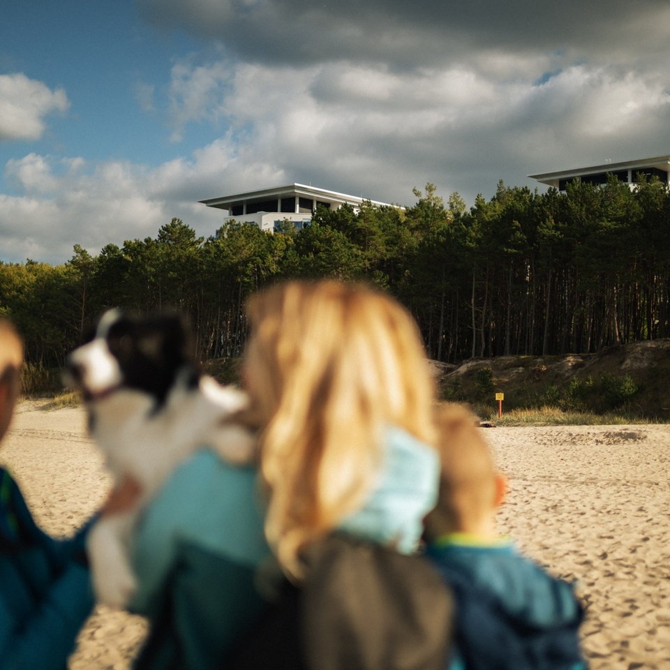 Famille avec chien sur plage de sable face à des bâtiments modernes au-dessus d'une forêt de pins sous un ciel nuageux