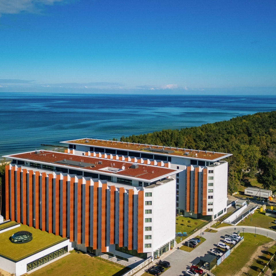 Nowoczesny hotel przy plaży, dwie bryły z pionowymi drewnianymi panelami, zielone tarasy dachowe, parking, widok na sosnowy las i morze