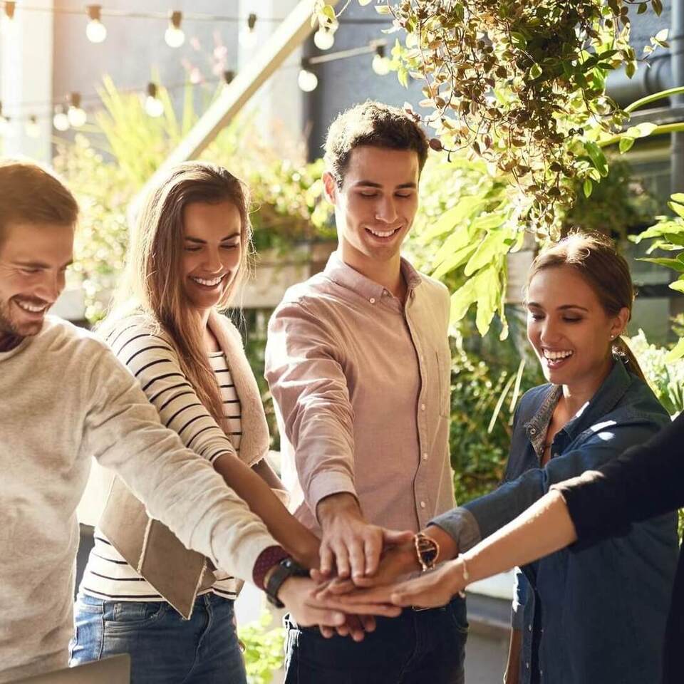 Freunde legen Hände zusammen auf sonniger Hotelterrasse mit Pflanzen und Lichterketten, entspanntes Treffen