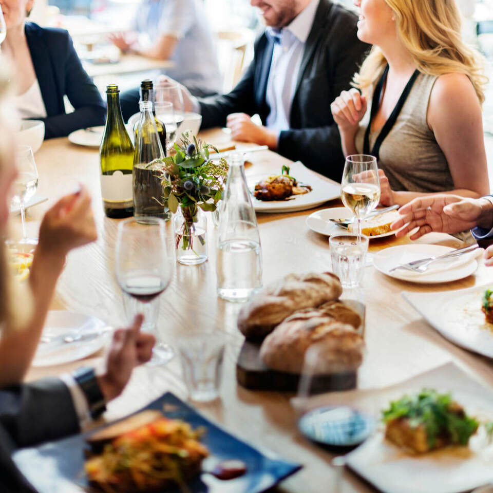 Geschäftsgäste an einem Gemeinschaftstisch im Hotelrestaurant, Gourmetgerichte, Wein, frisches Brot und helle Tageslichtatmosphäre